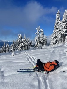 Maiya lays back on her skis and pack on the ski tracks. Her orange jacket stands out in the white snow and blue sky overhead. 